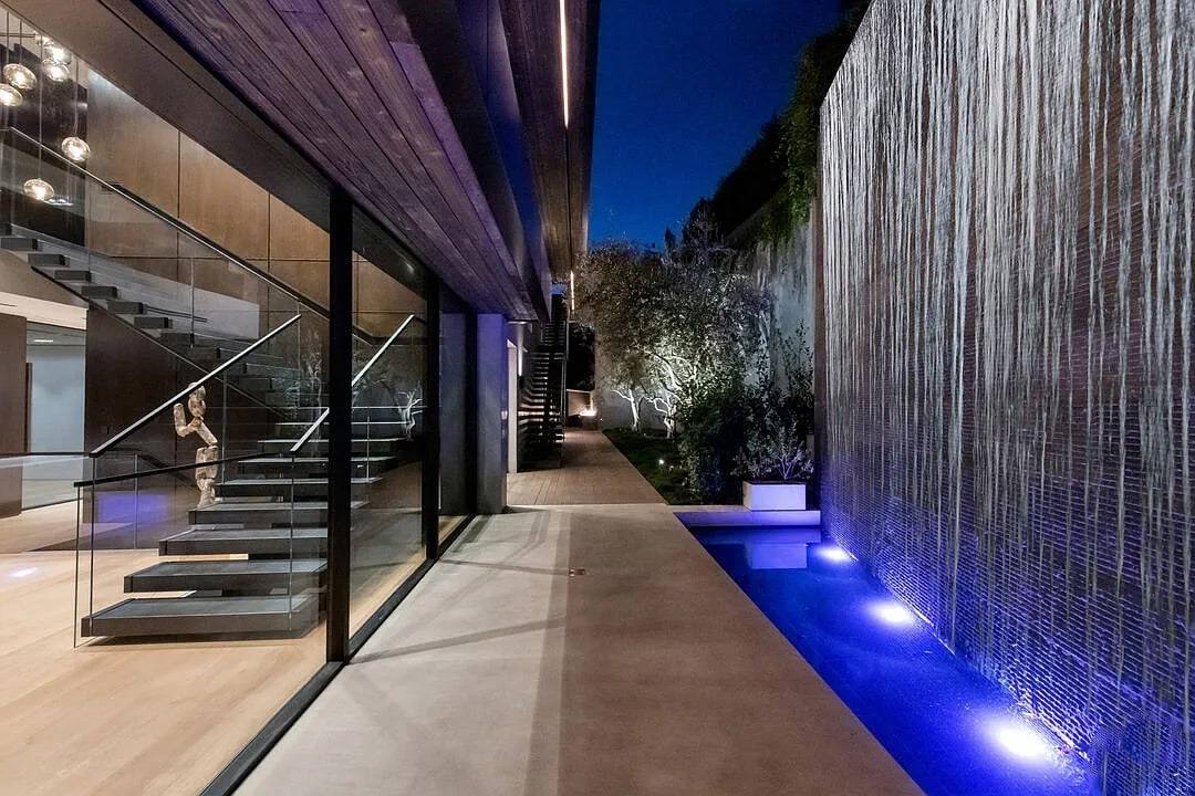 Night view of a glass-walled stair and a lit water wall beside a narrow pool