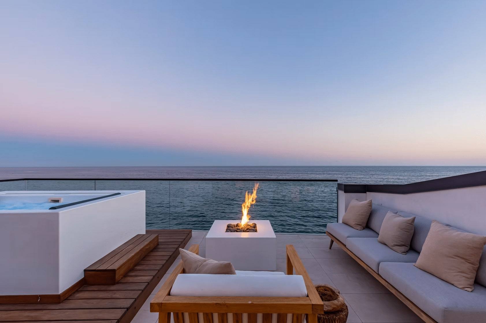 Rooftop deck of the Malibu Road residence at dusk, with a fire bowl, built-in seating and the Pacific beyond the glass rail