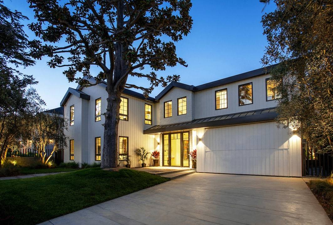 White two-storey Beverly Hills residence at dusk, with a dark metal roof, lit windows and a mature tree over the drive