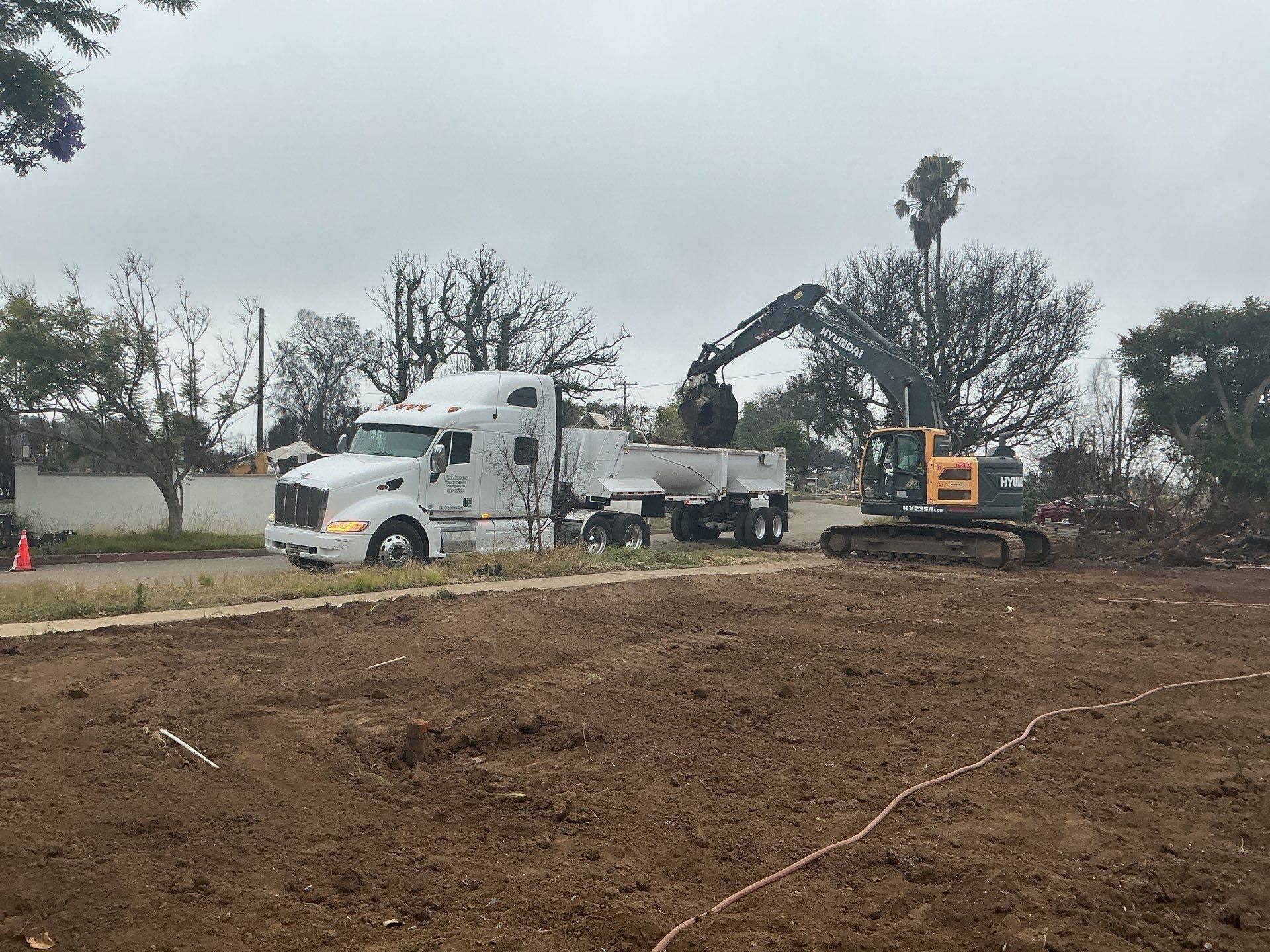 Excavator loading a dump truck at the edge of a cleared Pacific Palisades lot