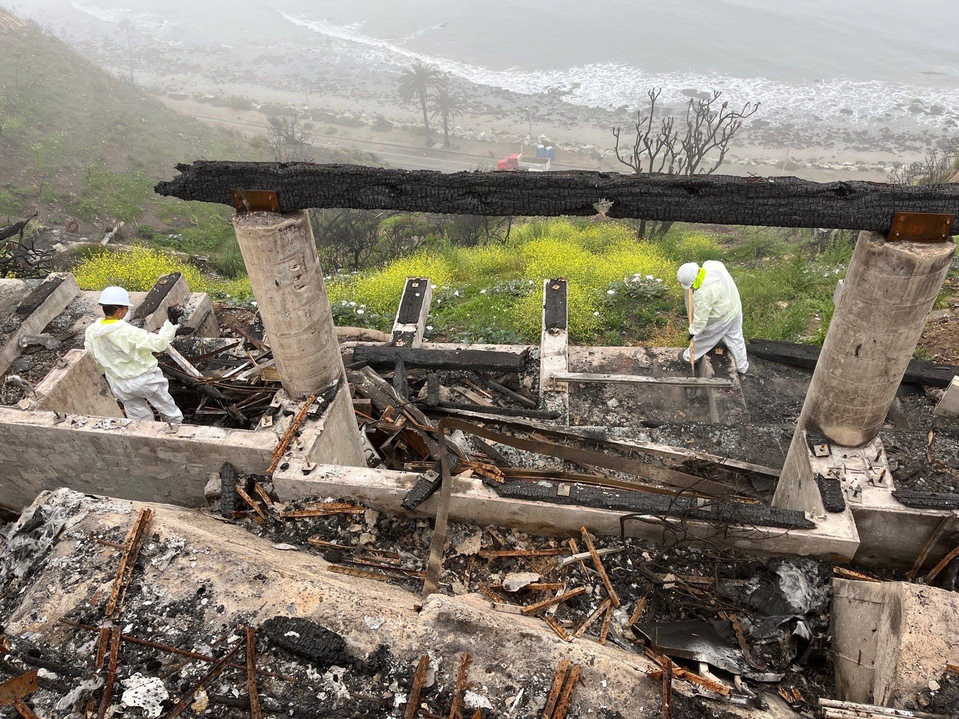 Crew in protective suits removing burned timber on a Malibu hillside lot above the ocean
