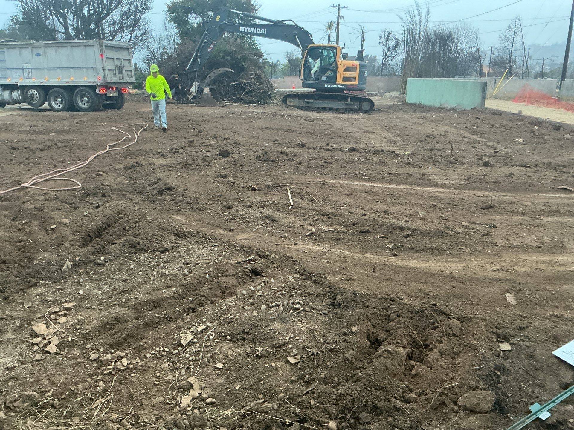 Crew member watering the cleared lot while an excavator and a dump truck work behind