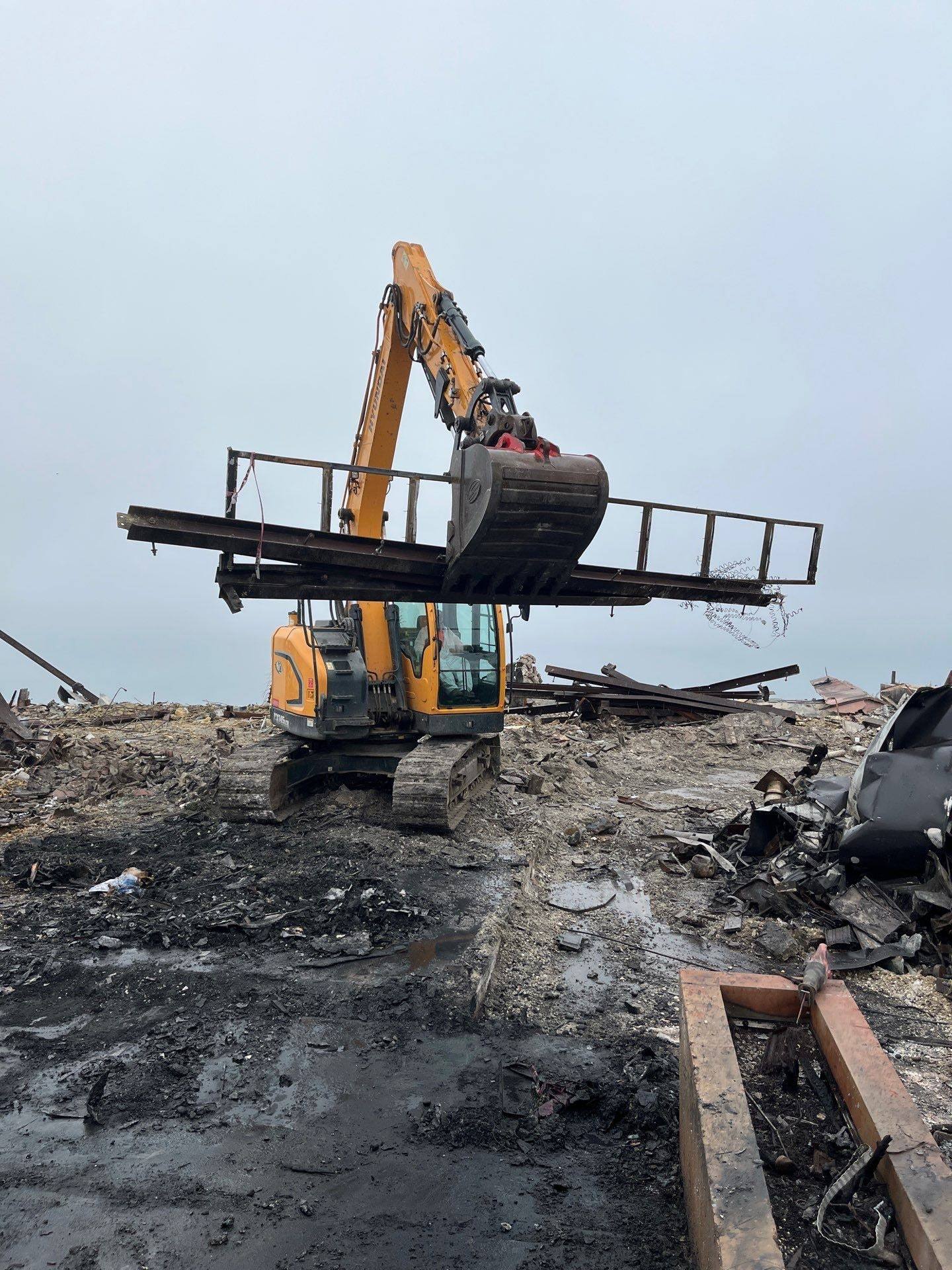 Excavator lifting a burned steel frame against the ocean haze