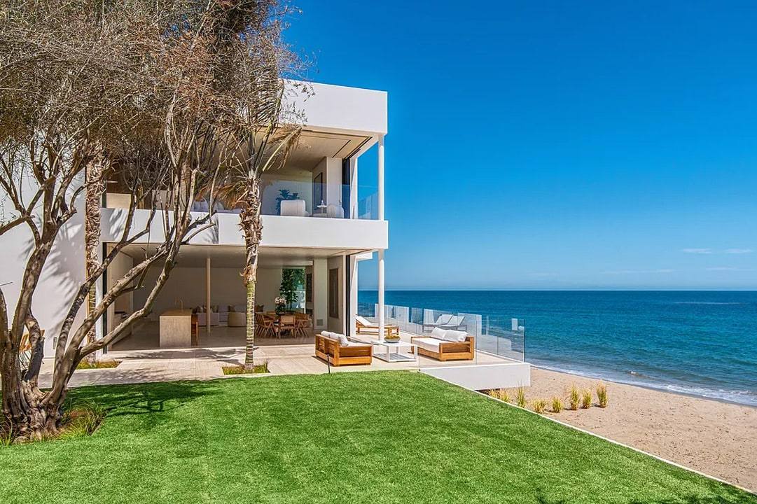 Two-storey white beach house with glass balconies above a lawn that runs to the sand