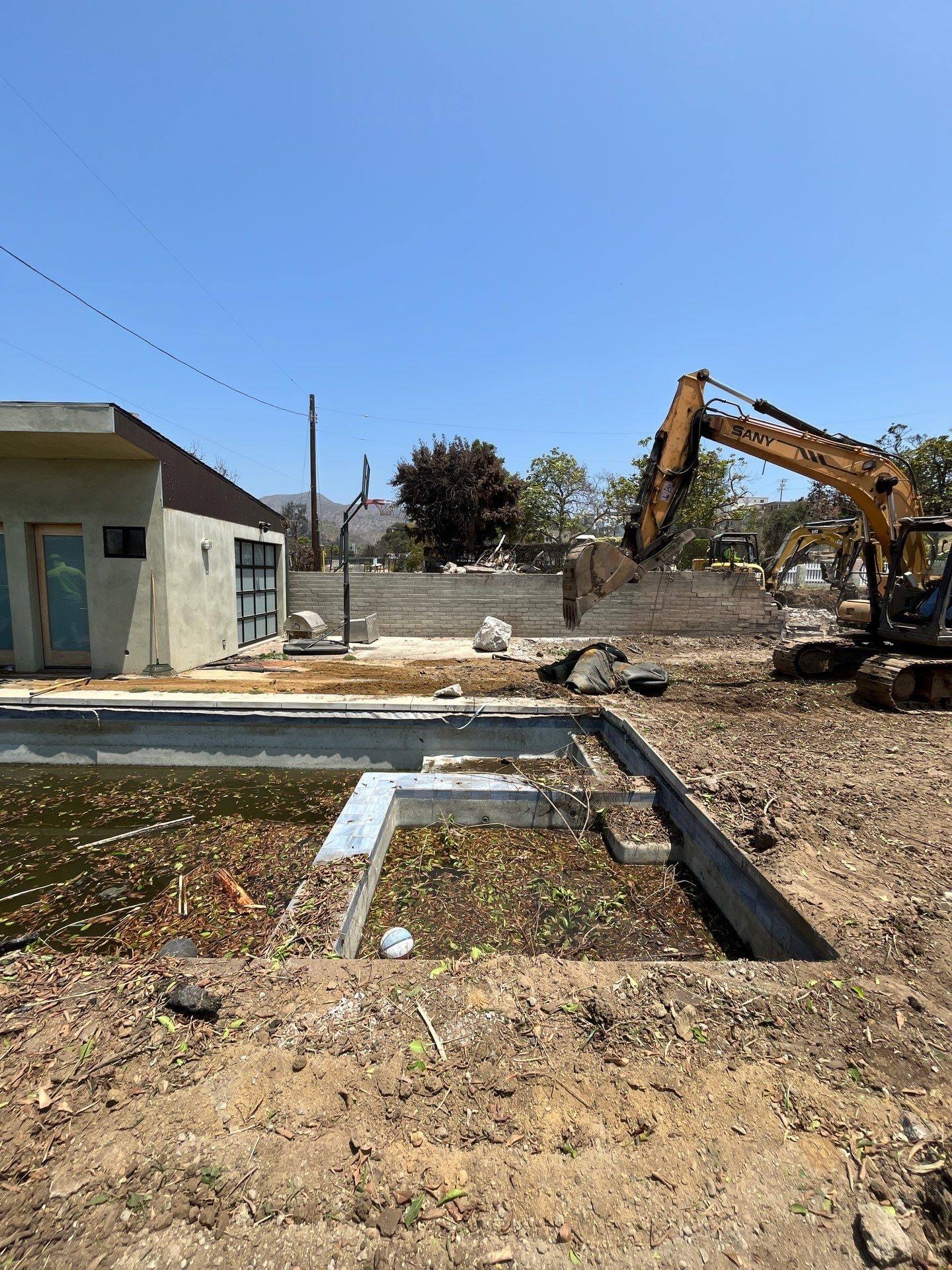 Excavator working past a remaining pool shell and a small outbuilding